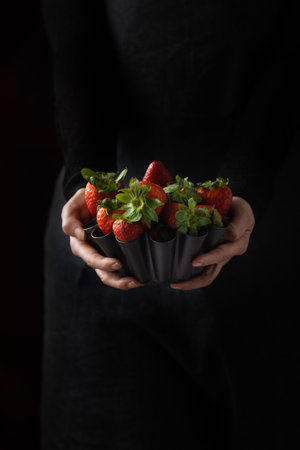A woman holding a metallic bowl filled with ripe, red strawberries ready to eat, set against a black backgroundの写真素材