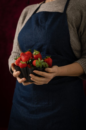 A woman holding a metallic bowl filled with ripe, red strawberries ready to eat, set against a black backgroundの写真素材