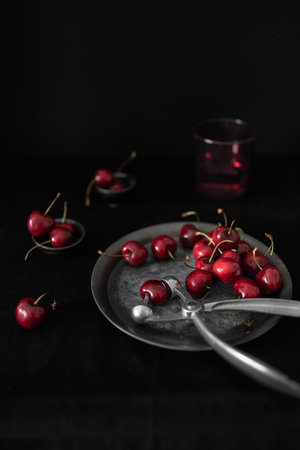A plate of fresh cherries accompanied by a cherry pitter, showing the preparation process against a dark backgroundの写真素材