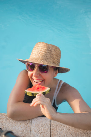 A smiling woman wearing sunglasses and a straw hat, savoring a slice of watermelon while relaxing in a swimming poolの写真素材