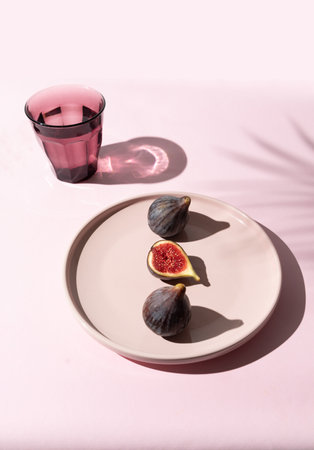 A purple fig cut in half next to two whole figs on a pink plate against a soft pink background....
