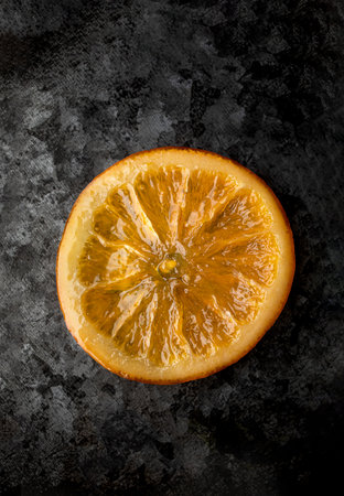 A single glossy orange slice, perfectly caramelized, placed on a textured dark metallic surface, showcasing intricate details and vibrant colorsの写真素材