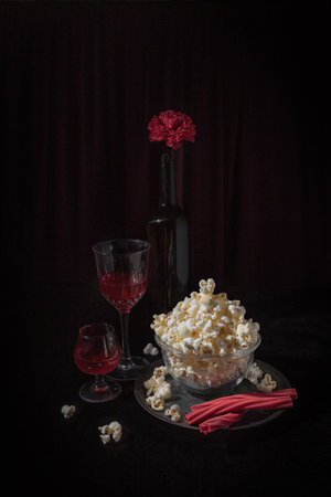 A cinematic-inspired still life featuring a bowl of popcorn, red drinks in elegant glasses, and a bottle with a red carnation on top. The setup evokes the ambiance of a cozy weekend movie night, with a dramatic dark background that mimics a theater-like atmosphereの写真素材