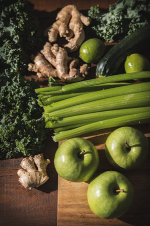 Fresh green detox ingredients like celery, kale, green apples, ginger, and lime arranged on a wooden surfaceの写真素材