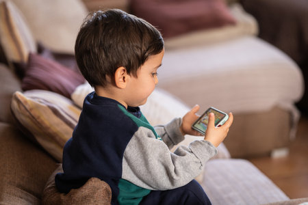 A young boy sitting on a couch, holding a smartphone with fascination, highlighting kids' early...
