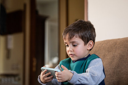 A three-year-old child sitting on a couch, looking at a smartphone, highlighting modern childhood...
