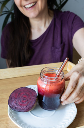 A fresh beetroot smoothie sits in the foreground while a woman in the background reaches to drink it. The image captures the concept of wellness, natural nutrition, and a healthy daily routine through plant-based habits.の写真素材