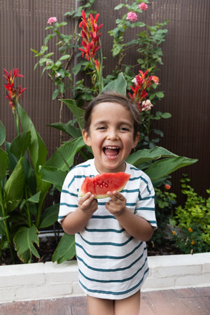 A cheerful child in a striped shirt laughs out loud while enjoying a slice of juicy red watermelon.の写真素材