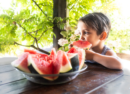 A cheerful child takes a big bite of a slice of juicy red watermelon while sitting at a garden table, surrounded by lush green plants.の写真素材