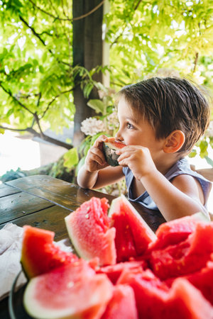 A cheerful child takes a big bite of a slice of juicy red watermelon while sitting at a garden table, surrounded by lush green plants.の写真素材