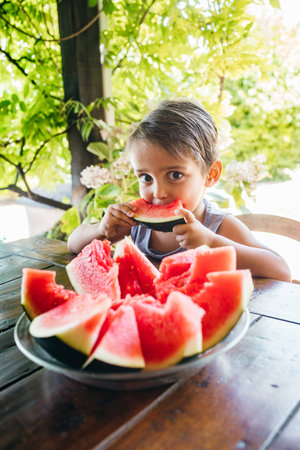 A cheerful child takes a big bite of a slice of juicy red watermelon while sitting at a garden table, surrounded by lush green plants.の写真素材