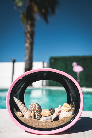 Close-up of pink diving goggles filled with beach sand and seashells, placed with a swimming pool in...