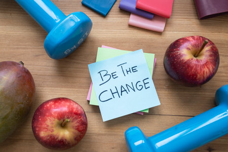 Green sticky note with handwritten âBe the Changeâ message placed on a table surrounded by colorful fruits and workout accessories, symbolizing motivation, sustainability, and a healthy, mindful lifestyle.の写真素材
