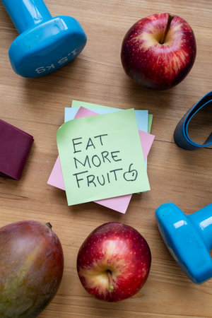 Green sticky note with handwritten âEat More Fruitâ message placed on a table surrounded by colorful fresh fruit and workout accessories, promoting healthy eating, wellness, and active lifestyle.の写真素材