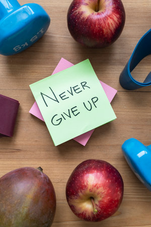 Green sticky note with handwritten âNever Give Upâ message placed on a table surrounded by fresh fruit and sports accessories, conveying motivation, resilience, and a healthy active lifestyle.の写真素材