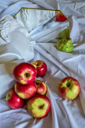 Ancient greek white sculpture plaster copy marble head of diana reflected in a mirror in gold frame against a background of white drapery fabric with folds with green pears and red yellow applesの写真素材