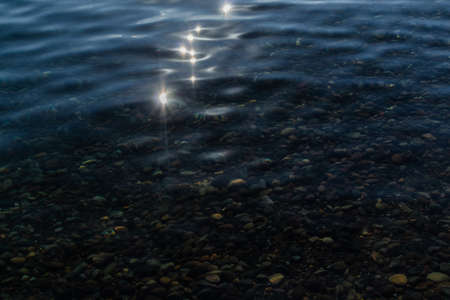 sun glare and green colorful pebbles in the clear blue water of lake baikal, ripples, shore, summerの写真素材