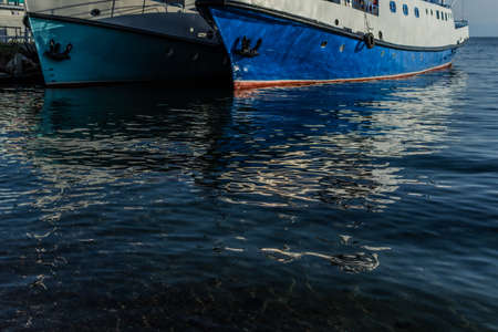 white blue ships boats bottoms on pier in bay of lake baikal with reflections in water in light of sunsetの写真素材