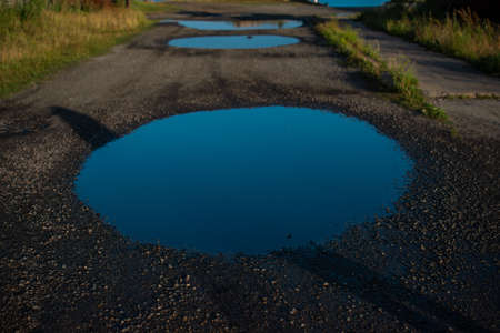 large geometrically smooth round blue water puddles with sky reflection in pebbles of gravel road with green grassの写真素材