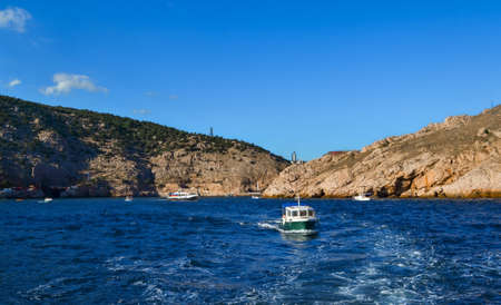 white yacht sails in blue sea among big red stones cliffs, mountains, summer light of sun, vacation
