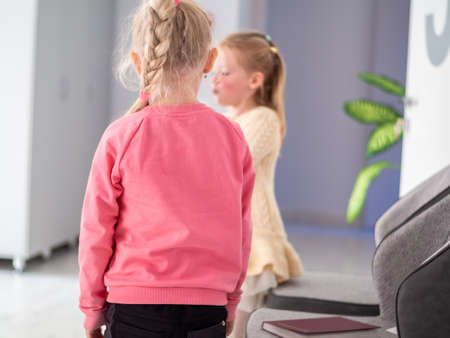 A charming girl in pink blouse and striped pants stands in front of another little girl in a...