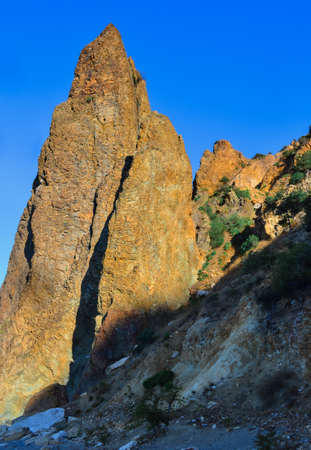 Yellow high sharp ancient basalt rock, cliff stands on Black Sea stones shore, in the light of sunset with shadows against the backdrop of blue summer sky, Crimeaの写真素材