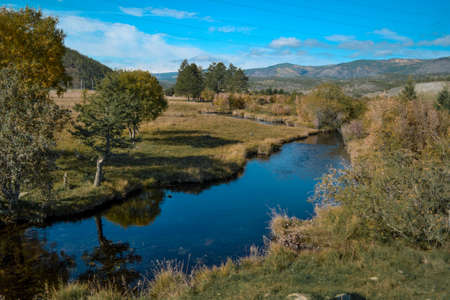 Bright blue winding river flows in grass, bushes, trees among the mountains and sky with clouds. Reflections in clear water. Autumn russia landscapeの写真素材