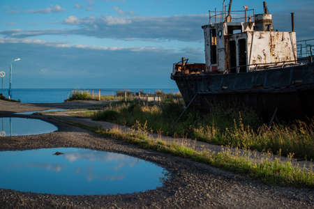 old rusty white ship, boat on grassy shore in light of sun, near round puddles on coast of lake...