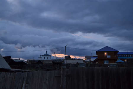 View of houses and church of village behind flat wooden Russian fence in evening. Cloudy blue sky,...