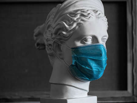 Plaster copy of the head of the ancient statue of Diana on a light textured back. The face of a woman with a plaster of Paris sculpture in a medical mask. Profile view. Covid-19 virus. Pandemicの写真素材
