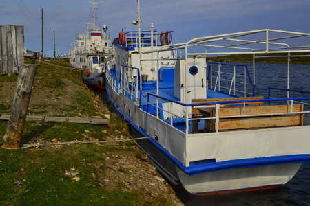 Large white blue boats tied to poles, ship stands by green grassy shore with wood rustic fence in...