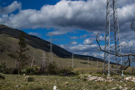 Power poles on yellow grassy steppe with green trees stand by slope of Baikal mountains. Metal...