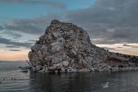 Stones rocky island with black birds, cormorants in the middle of Lake Baikal. Seascape with ripples, waves, sunset blue sky with clouds backgroundの写真素材