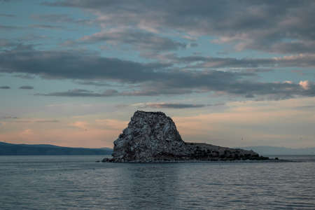 Stones rocky island with black birds, cormorants in the middle of Lake Baikal. Seascape with ripples, waves, sunset blue sky with clouds, mountains backgroundの写真素材