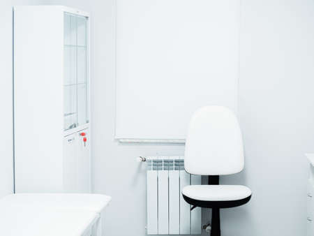 White black chair and medical couch for examining and treating a patient in light clean doctor's office. Window with curtain, cupboard, battery in the room. Workplaceの写真素材