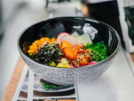 Poke bowl with radish, salmon, avocado, corn, seaweed, peas, nori, daikon, sesame seeds in black plate on blank menu, table in cafe, restaurant. Tasty natural foodの写真素材