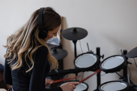 A beautiful woman in headphones sits at an electronic drum kit in the studio. Plays with drumsticks....