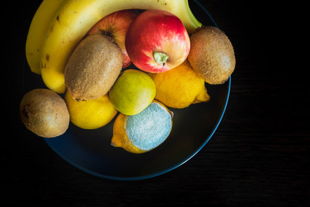 A dark blue plate with ripe colorful whole fruits: green and red apples, kiwi, yellow lemons, bananas. Concept spoiled lemon with white light textured mold. Top viewの写真素材