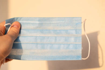 One disposable medical light blue mask in hand on light warm blurred background with shadow. Close-upの写真素材