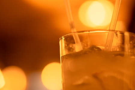 Rim of glass with lemonade, alcoholic cocktail, drink with ice cubes, lemon wedges and two straws. Blurred warm bokeh backgroundの写真素材