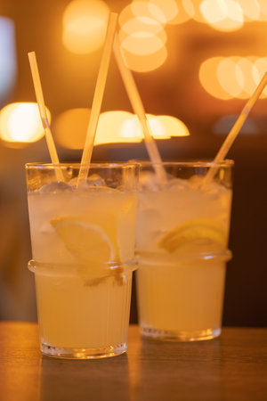 Two Lemonades in a glass with ice cubes on chopped lemon wedges. Refreshing cold drink with straws on the wooden table in a cafe. Blurred warm backgroundの写真素材