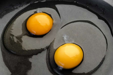 Two bright round yolks in raw whites, uncooked small fried eggs is a simple breakfast in a black skillet. Top view, close-upの写真素材