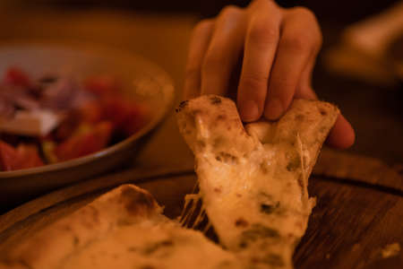 A hand takes a slice of hot four cheese pizza with stretched cheese close-up. Freshly prepared vegetarian Italian dish in warm evening lightの写真素材