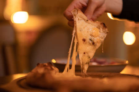 A woman's hand takes a slice of hot vegetarian four cheese pizza with stretched cheese close-up, warm evening light. Freshly prepared Italian dish in the restaurant, cafeの写真素材