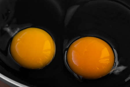 Two bright round raw yolk in raw white in a dark black frying pan. Cooking tender, soft fried eggs as a simple dish. Close-up, macroの写真素材