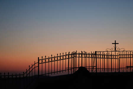 A fence with a religious cross attached to it next to a church and the aegean sea, while the sun is setting in a dramatic wayの写真素材