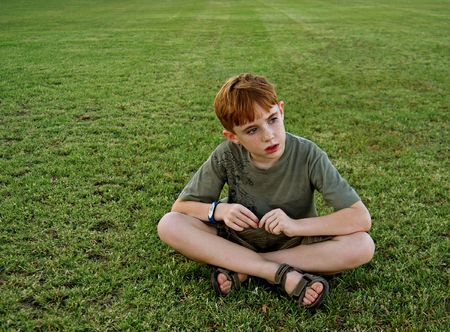 An 8-year-old boy sits on a grassy field and looks off out of frameの写真素材