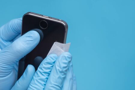 Hands in medical gloves wipe smartphone screen with disinfectant cloth. Close-up hands clean mobile phone on blue background. Closeup of a Doctor in gloves disinfecting his smartphoneの写真素材