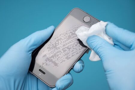 Closeup of a caucasian man disinfecting his smartphone. Hands in medical gloves wiping mobile phone with alcohol decontaminating agent, top side view on blue background. Copy spaceの写真素材
