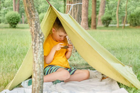 Boy playing video game outdoor in park, backyard, garden. Kid with phone in tent. 6 years old child having fun on nature. E-learning concept. Children and gadgets. Entertainment. Summer holidaysの写真素材